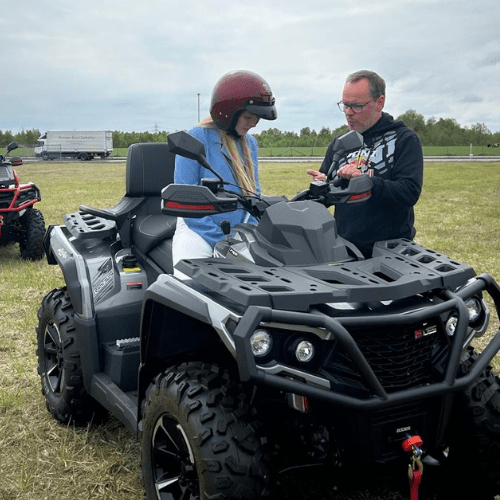 Prote Adventure Quad Vermietung Delmenhorst bei Bremen Prote Adventure Handel und Verleih von UTV ATV und Quads in Delmenhorst beo Bremen Woman wearing a red helmet sits on a dark gray ATV in a field while a man checks its controls nearby